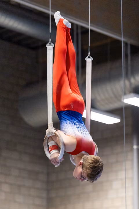 Gymnast performs inverted hang position on still rings, demonstrating strength and control in red and blue uniform