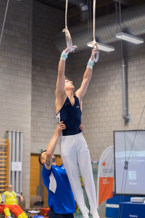 Male gymnast suspended from rings with arms extended upward while coach provides support from below in training facility