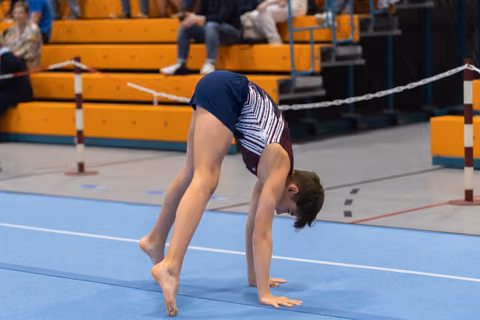 Young gymnast performs a bridge backbend on blue floor mat during gymnastics event with spectators in background