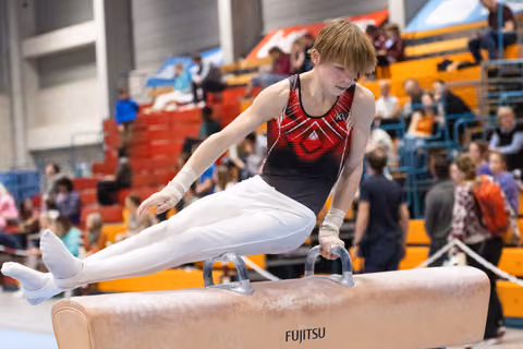 Young male gymnast performs scissors move on pommel horse, demonstrating strength and flexibility before crowd in gymnasium