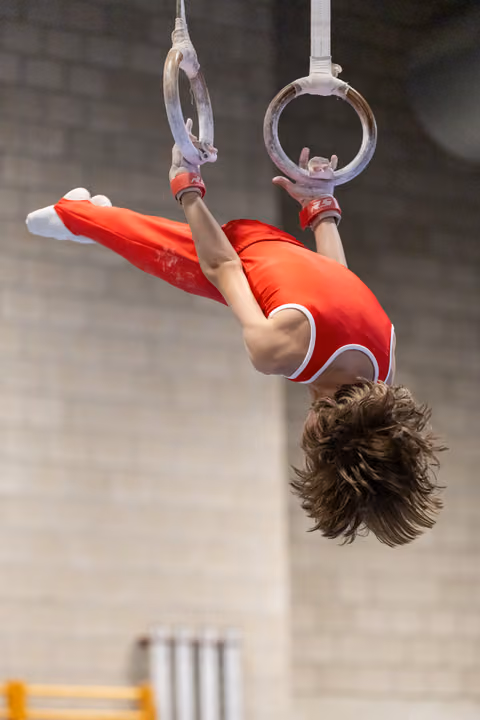 Gymnast performing an inverted hold on still rings, body horizontal in bright red uniform with white trim