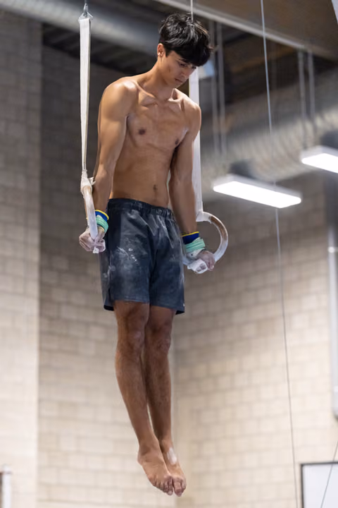 Shirtless gymnast performing still rings routine, showing intense concentration while suspended in gym training facility