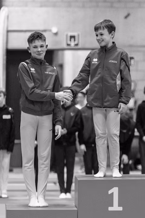 Two young gymnasts shake hands on podium, first and second place, both smiling in matching team jackets