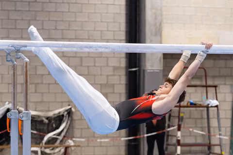 Gymnast in red and black leotard performs horizontal bar routine with extended body position and white pants on legs