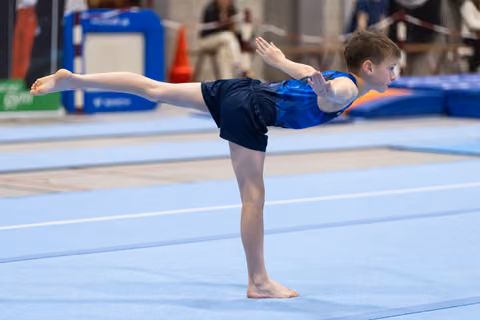 Young gymnast performs an arabesque balance with extended arms during floor exercise routine on blue training mat