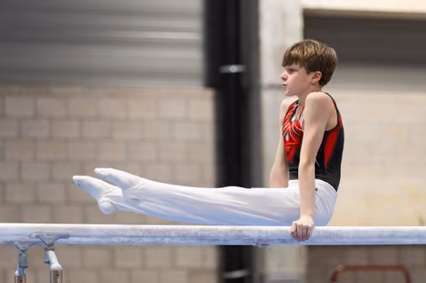 Young male gymnast holds an L-position on horizontal bar, legs extended, focused expression, in indoor gym facility