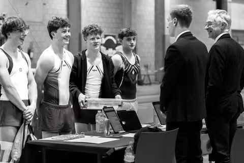 Four young gymnasts in competition attire stand smiling at a table, conversing with two officials in dark suits