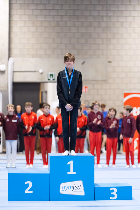 Young male gymnast stands proudly on first place podium wearing blue medal, while teammates in red and burgundy applaud behind him