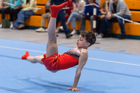 Young gymnast performs dynamic breakdancing freeze move on blue floor mat with spectators watching from bleachers