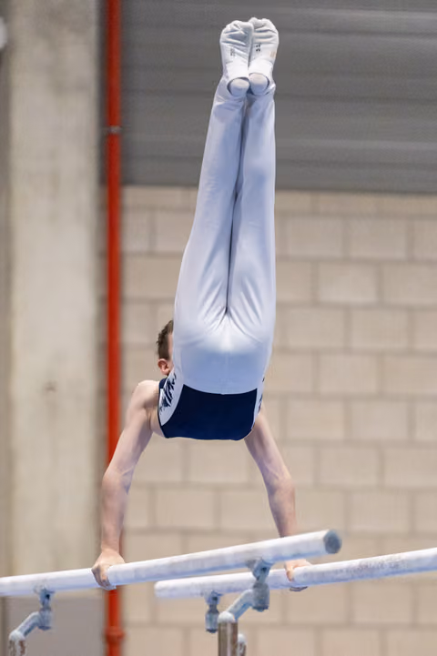 Male gymnast performs inverted handstand on parallel bars, body fully extended upward in white and blue uniform