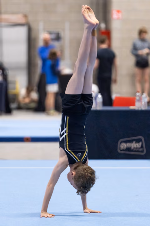 Young gymnast performs a handstand on blue floor mat during routine, legs extended straight up in competitive gym setting
