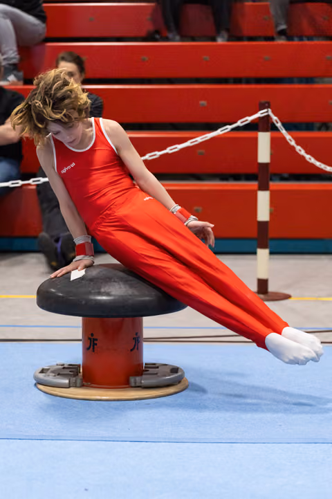 Gymnast in red uniform performing pommel horse routine with focused expression, body extended horizontally in gym
