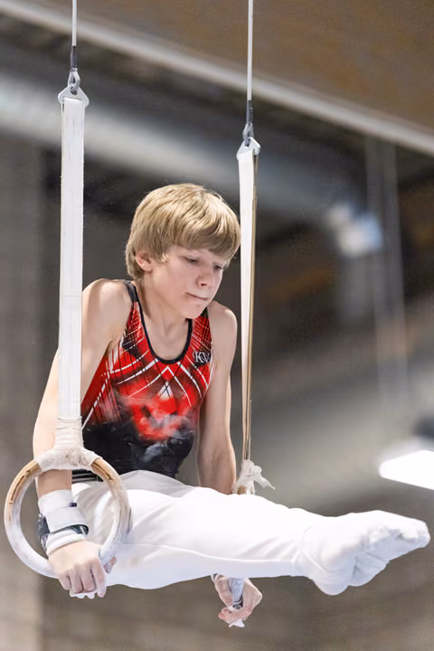 Young male gymnast performs on rings, concentrating intently on his form in white pants and red leotard
