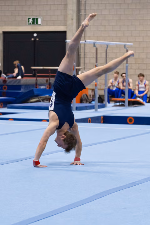 Male gymnast executing a one-armed handstand during floor routine, demonstrating strength and balance on blue mat