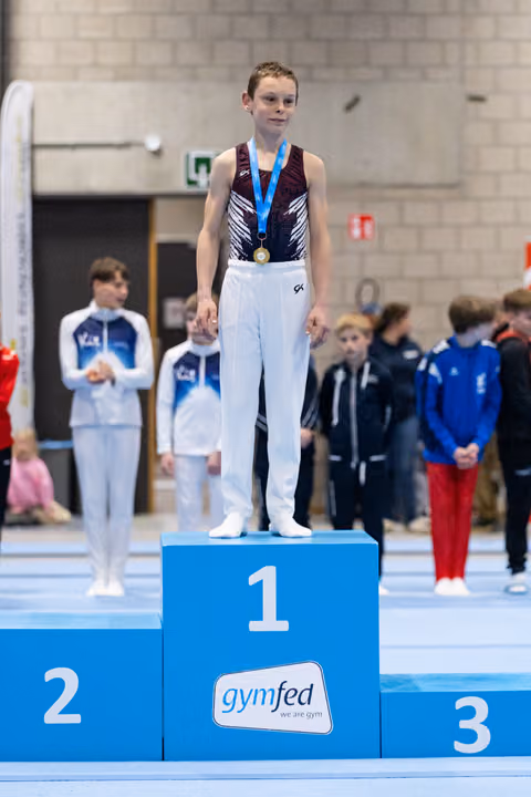 Young gymnast in maroon leotard stands on first-place podium wearing gold medal and blue ribbon at gymnastics meet