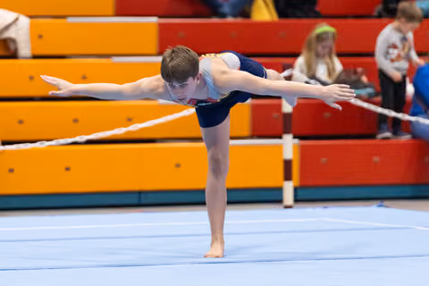 Male gymnast performs an arabesque balance on floor exercise, arms extended, spectators visible on colorful bleachers behind