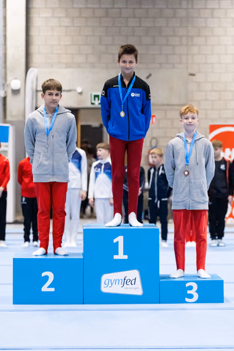 Three young gymnasts stand on award podium, winner in blue jacket on first place, two others in grey hoodies on second and third