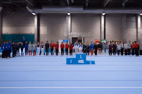 Multiple gymnastics teams standing on award podiums in an indoor sports hall during a medal ceremony celebration