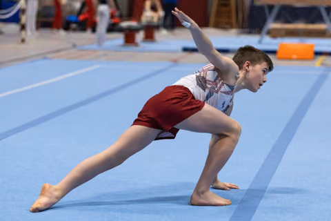 Young gymnast performs a lunge position during floor routine, arms extended with focused expression on blue mat