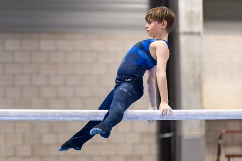 Young male gymnast performing a horizontal bar routine in blue and black competition attire, demonstrating strength and focus