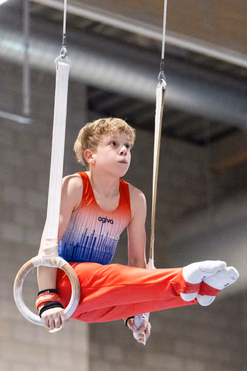 Young gymnast in red and blue gradient leotard demonstrates strength hold on rings, looking upward with focused expression