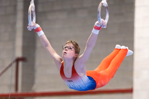 Young gymnast in orange pants and gradient leotard performs rings routine with extended body position, wearing glasses