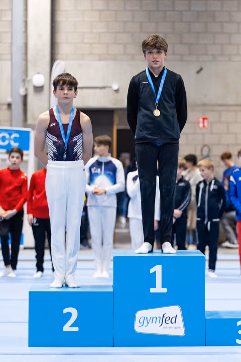 Two young gymnasts stand on award podium wearing medals, first place winner elevated above second place finisher