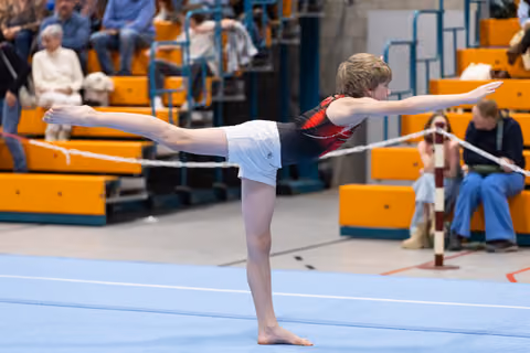 Young gymnast performs arabesque balance during floor exercise routine while spectators watch from orange bleachers