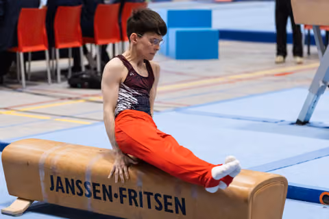 Male gymnast in sequined leotard and red pants sits on pommel horse, focused and preparing for his routine at indoor meet