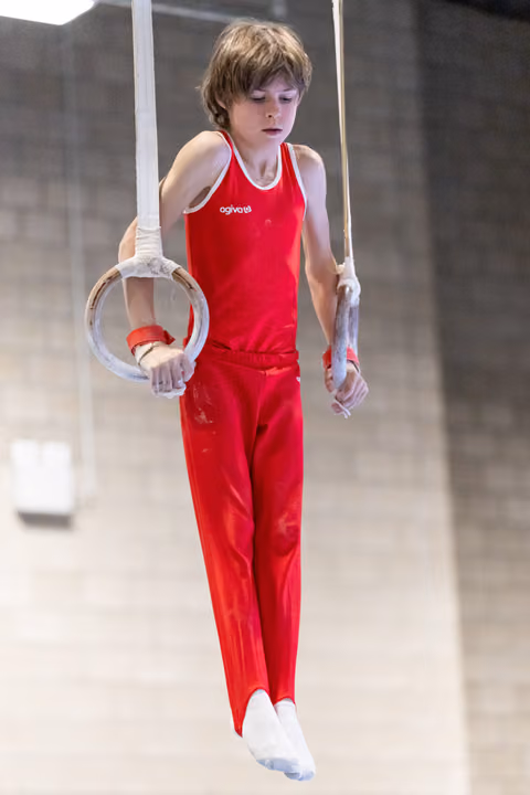 Young gymnast in red uniform performs on rings with chalk-dusted hands, displaying intense concentration during routine
