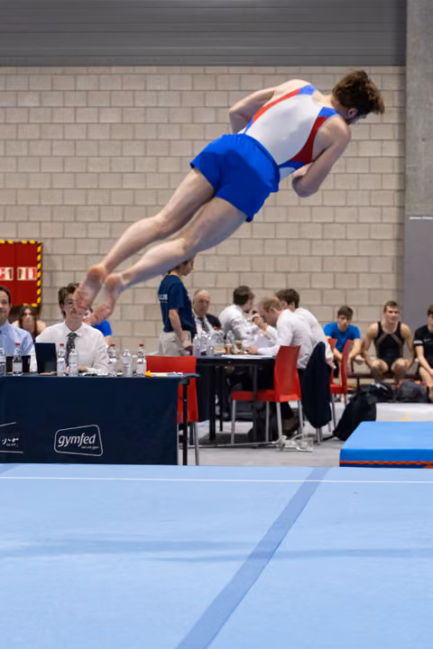 Male gymnast in horizontal flight during floor routine, wearing red and blue uniform, with judges and spectators in background