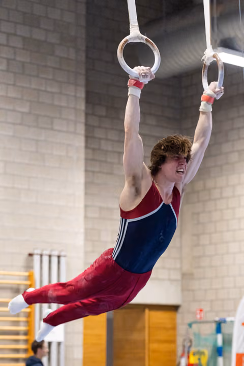 Young male gymnast performing on rings with extended body position, wearing red and blue leotard in indoor training facility
