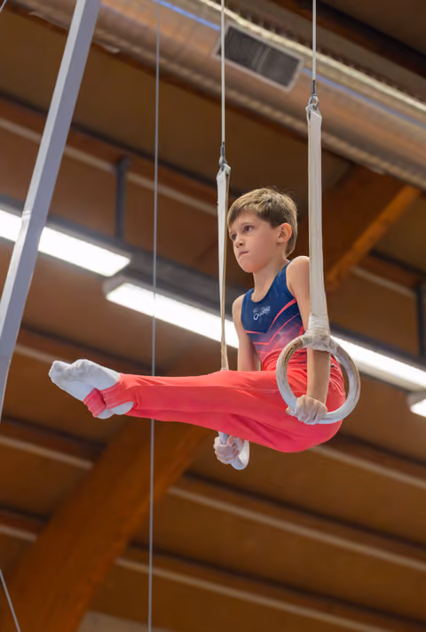 Determined young boy holds gymnastics rings in a split position, legs extended, concentrated gaze in an indoor gym.