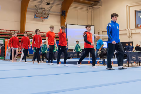 Young male gymnasts in red and blue jackets march across the floor exercise mat during a gymnastics competition.