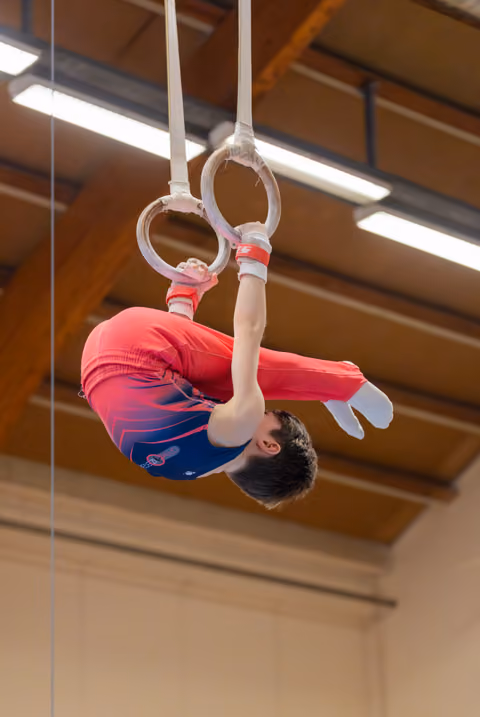 Young male gymnast performs an inverted tuck on still rings, wearing a red and blue leotard in an indoor gymnasium.
