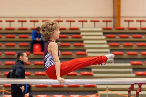 Young gymnast holds a precise L-sit on parallel bars, legs extended forward, focused expression in a nearly empty red-seated arena.