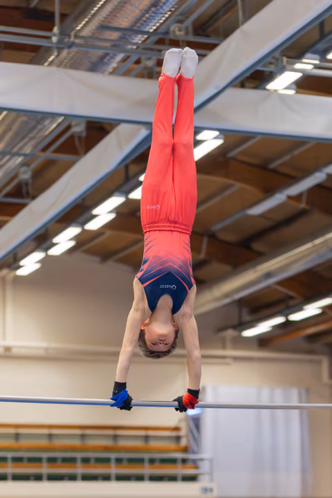 Young male gymnast performs an inverted handstand on the high bar, body fully extended, in an indoor gymnastics hall.