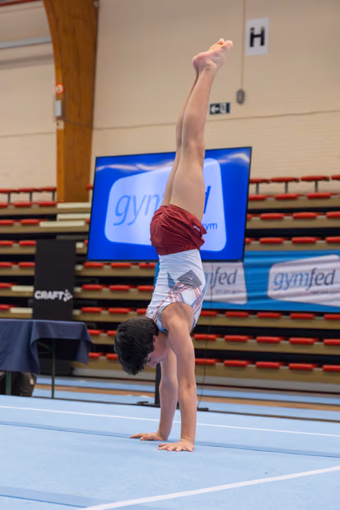 Young gymnast performs a handstand with legs split on a blue floor mat at a Gymfed competition.
