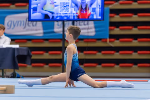 Young male gymnast performs a perfect split on the floor, back arched, focused expression during an indoor gymnastics meet.