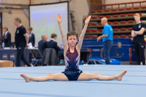Young gymnast performs a split on the floor with arms raised high, focused expression during a gymnastics meet.