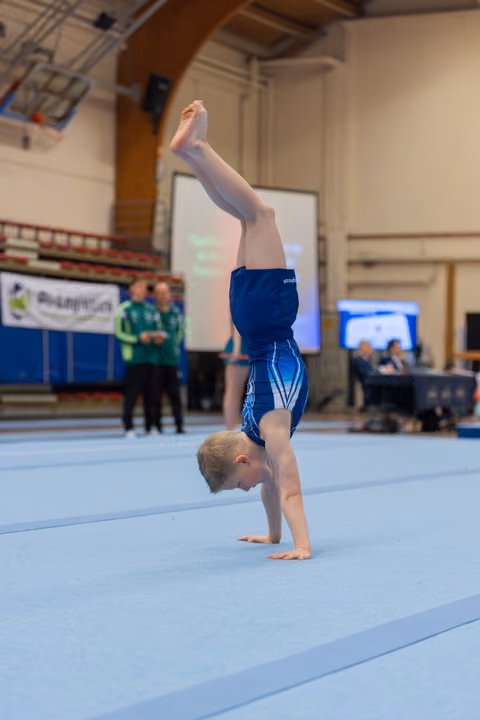 Young male gymnast performing a precise handstand on a blue mat during an indoor gymnastics competition.
