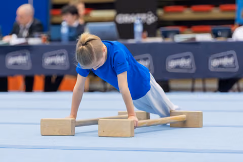 Young child in blue shirt performs parallel bar exercise on blue mat, leaning forward with intense concentration.