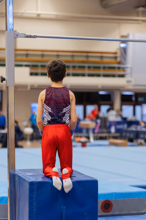 Young male gymnast stands on a blue block, back turned, gazing at the high bar ahead with quiet focus in a gym.