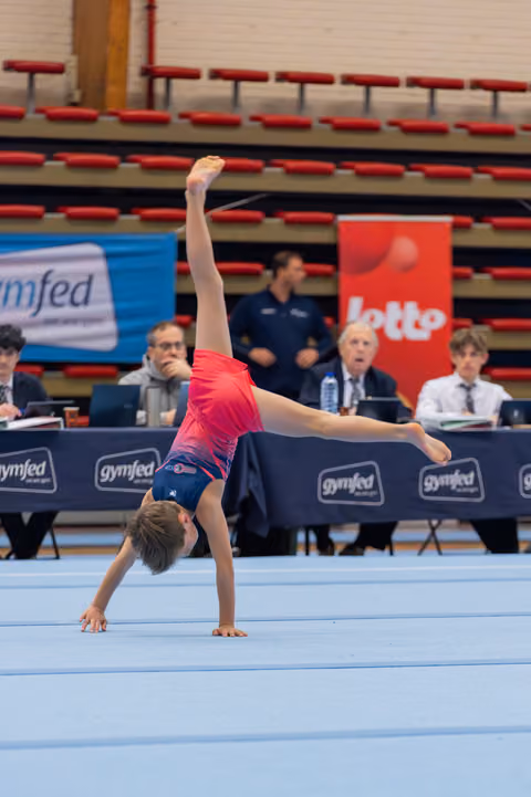 Young gymnast performs a cartwheel on the floor exercise mat, judges observing at the scoring table behind.