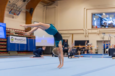Young gymnast performs a handstand on the floor exercise mat, body arched with legs extended, in a gymnastics hall.