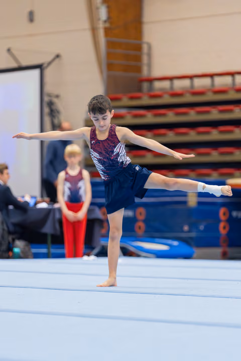 Young male gymnast balances on one leg with arms outstretched during a floor routine at an indoor gymnastics meet.