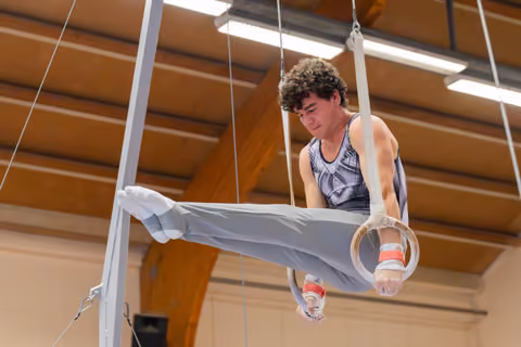 Young male gymnast with curly hair performs a straddle hold on rings, focused expression, in a wooden-ceiling gymnasium.