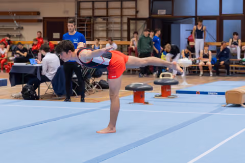 Young male gymnast holds a precise arabesque balance on the blue floor mat during an indoor gymnastics competition.