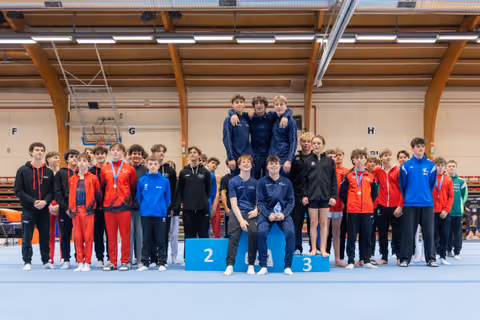 Young male gymnasts pose on podium, smiling proudly; teammates in red, black and blue surround them in a sports hall.