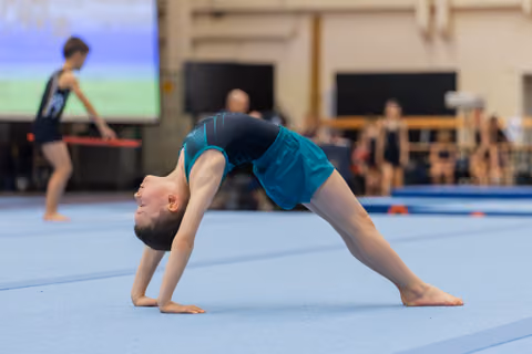 Young gymnast performs a bridge pose on the blue floor mat, head dropped back, focused expression during a gymnastics event.